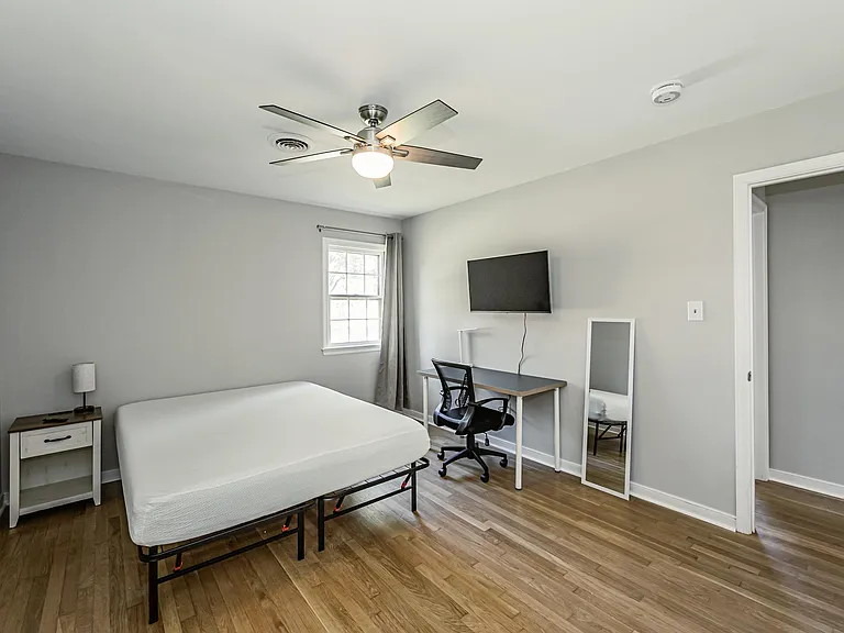Well-proportioned bedroom with a ceiling fan, hardwood-style floors, and a work desk plus wall-mounted TV and mirror.