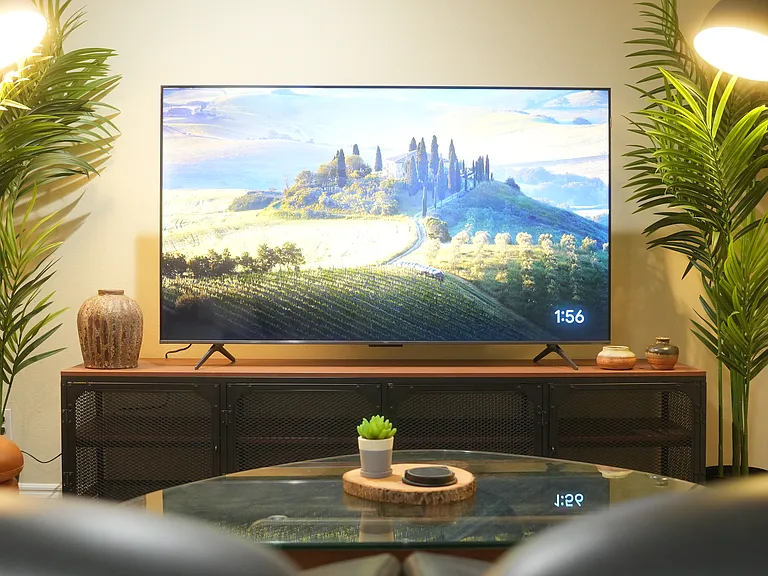 The living room has 2 black leather arm chairs and a tan leather ottoman along with a 65" inch smart TV and an Herman Miller inspired glass coffee table