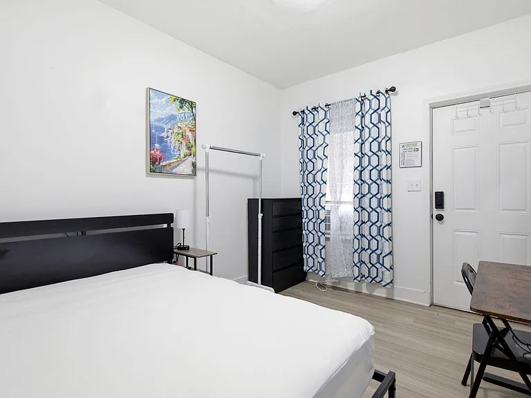 Private bedroom with a full bed, dresser, nightstand, and work desk, plus a window with curtains and A/C unit.