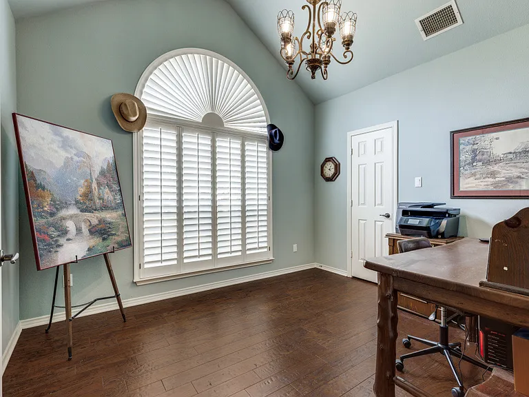 This bedoom (can be used as an office) fetures a chandelier, dark wood-type flooring, and vaulted ceiling. Plantation shutters cover large wnidow.