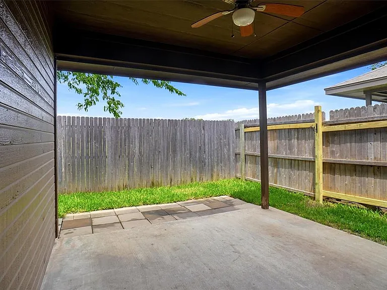 Covered concrete patio featuring a ceiling fan, a slatted exterior wall, and a wooden privacy fence enclosing a grass area