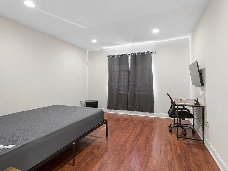 Spacious bedroom with hardwood floors, recessed lighting, window coverings, and a dedicated desk area with wall-mounted TV.