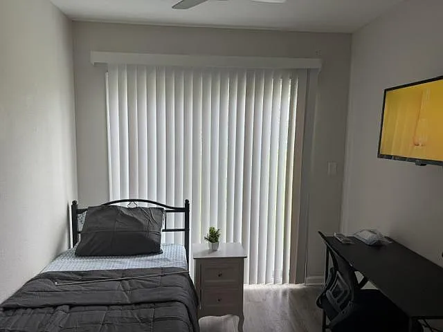 Private bedroom with a furnished twin bed, desk workspace, and large sliding door with vertical blinds for natural light.