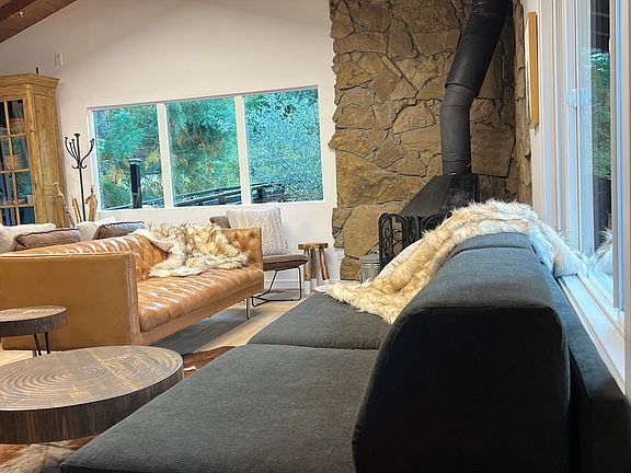 Cozy seating area with a stone fireplace and large windows framing forest views. Warm wood beams and rustic decor create the perfect spot to relax, unwind, and enjoy the Big Bear cabin atmosphere.