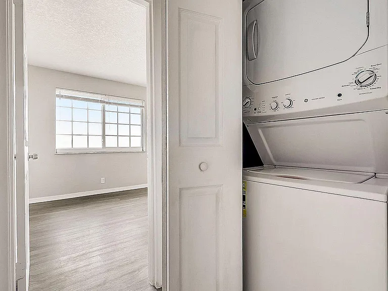 Bright apartment with modern in-unit stacked washer and dryer adjacent to spacious sunlit bedroom featuring large window views.