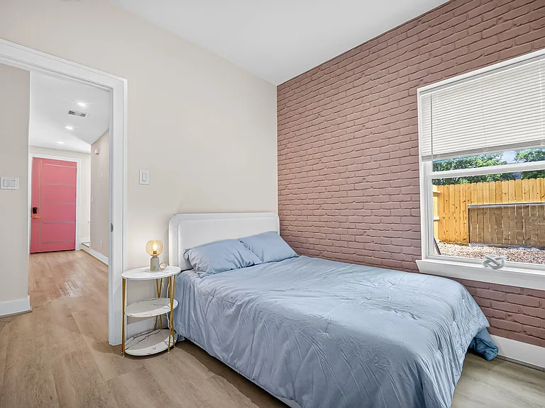 Bedroom features a comfortable bed, accent brick wall, and a large window with blinds for natural light and privacy.