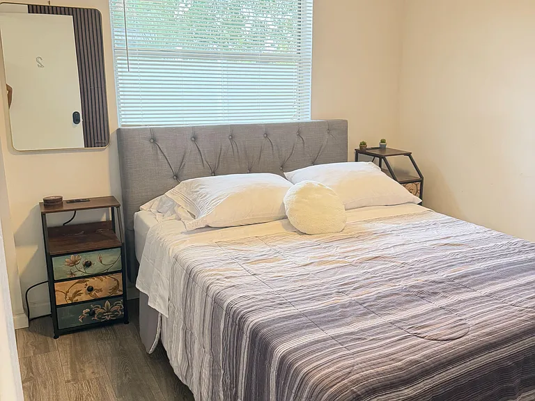 Furnished bedroom with a full bed, bedside tables, and a large window with blinds for comfortable light control.