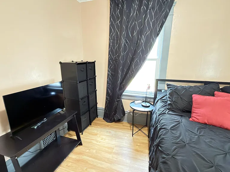Furnished bedroom with a bed, TV stand, and convenient storage, plus a window with curtains and hardwood flooring.