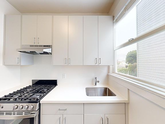 Bright kitchen with cabinets, gas stove, and stainless steel sink. Large window on the right provides natural light, creating a clean, airy feel.