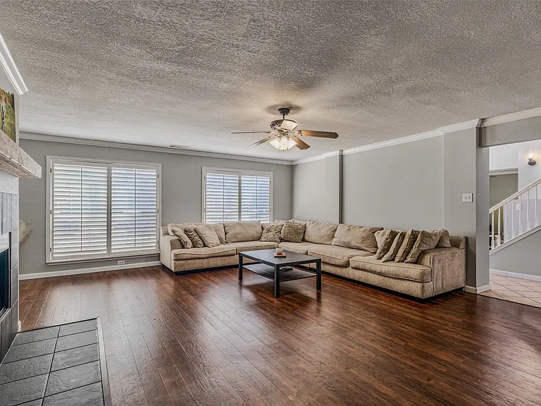 Spacious living area featuring wood-finish flooring, a tiled fireplace with wood mantel, and multiple windows equipped with plantation shutters