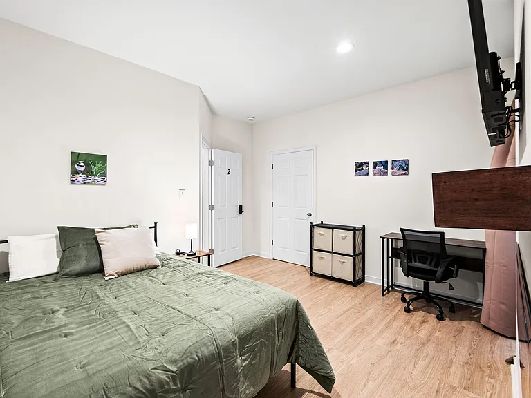 Furnished bedroom with full bed, wall-mounted TV, closet, dedicated desk workspace, and extra storage on wood-look floors.