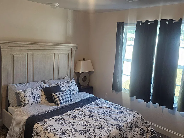 Furnished bedroom with a queen bed, nightstand and lamp, plus a large window with curtains for adjustable light.
