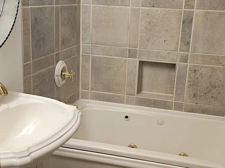 Beautiful full bathroom featuring luxurious Italian porcelain tile, a deep soaking tub with shower, and premium Kohler fixtures. The space includes a classic pedestal sink, round mirror, and elegant lighting