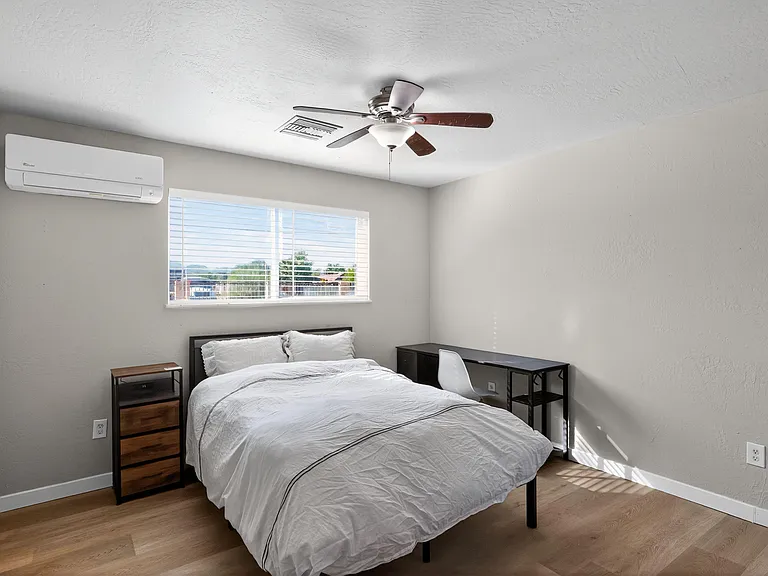 Furnished bedroom with a full bed, nightstand, and work desk, plus a large window and ceiling fan for everyday comfort.