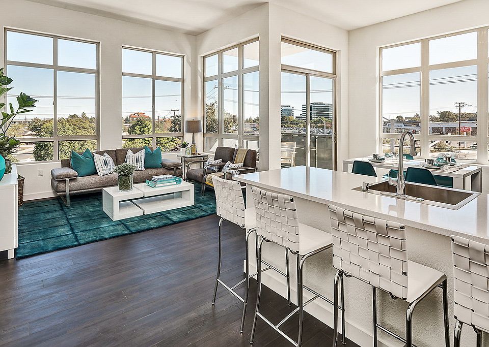 Staged, open living room and kitchen with hardwood-style, vinyl flooring and floor to ceiling windows.