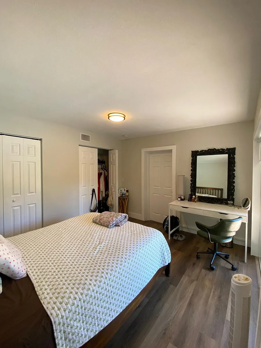 Spacious bedroom with a full bed, wide plank flooring, and a dedicated desk/vanity area plus closet storage. - NE 26th Ave