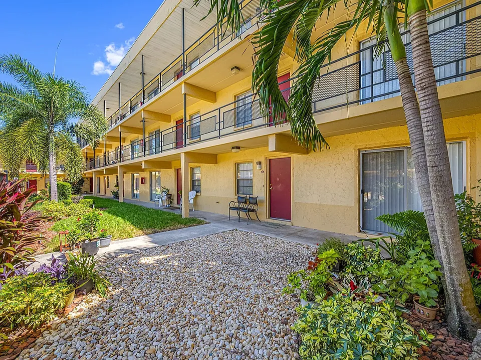 Charming two-story apartment community with vibrant garden landscaping and palm trees, set under a clear blue sky.
