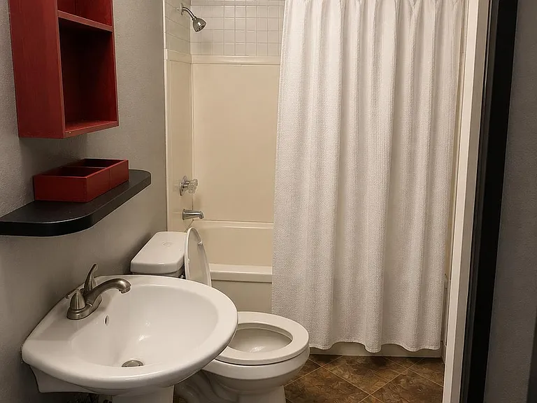 Full bathroom with tub/shower combo, tile surround, pedestal sink, and open wall shelving for everyday storage.