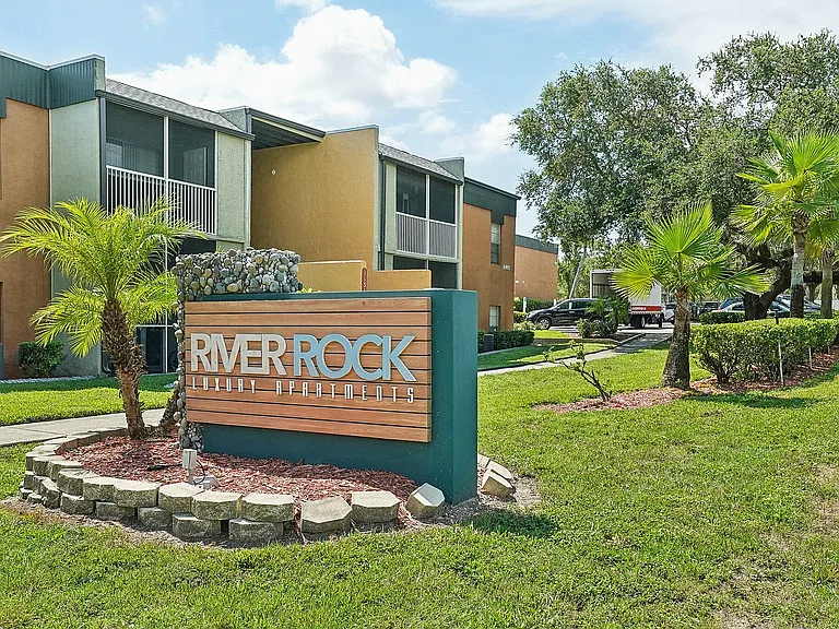 Inviting entrance to River Rock Luxury Apartments with lush greenery and modern signage.