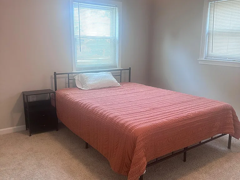 Furnished bedroom with a full-size bed, side table, and two windows with blinds for natural light and privacy.