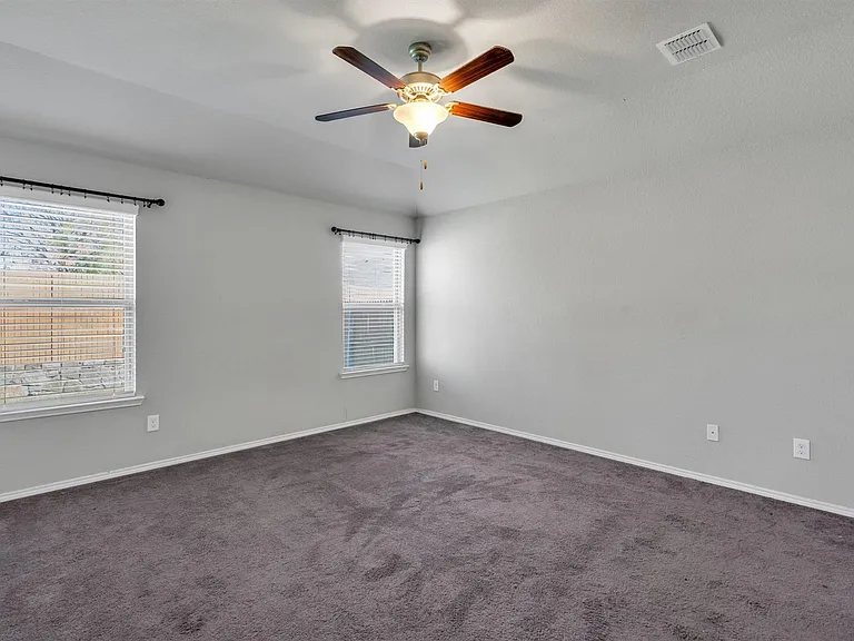 Spacious room featuring neutral paint, gray carpeting, dual windows with blinds, a ceiling fan with integrated lighting, and white baseboards