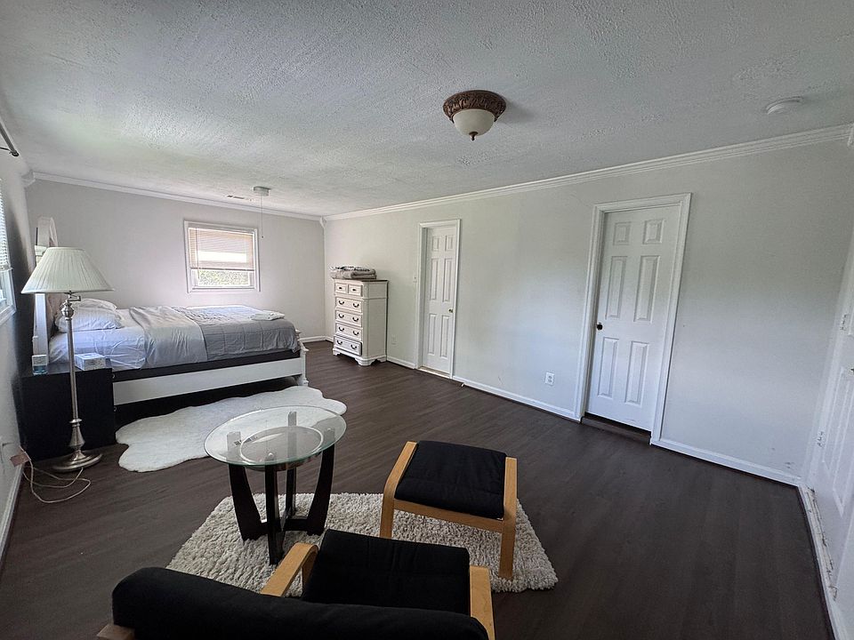 Spacious furnished bedroom with wood-style flooring, a comfortable bed, dresser storage, and windows with blinds.