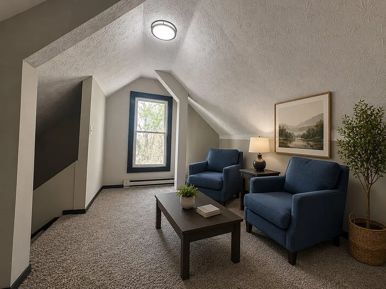 Finished loft-style sitting area with carpeted floors, dormer window, and space for two chairs plus a side table.