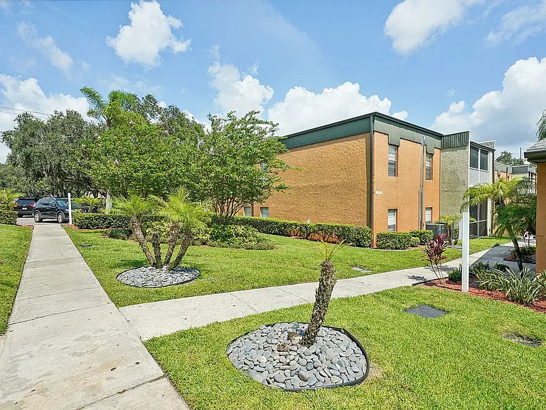 Charming apartment complex with well-manicured lawns, palm trees, and a pathway leading to the entrance under a blue sky.