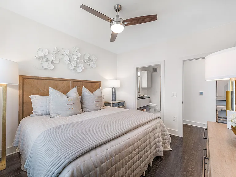 Bright and elegant bedroom featuring a plush king-sized bed with stylish decorative pillows, modern ceiling fan, chic wall art, and a glimpse into the en-suite bathroom.