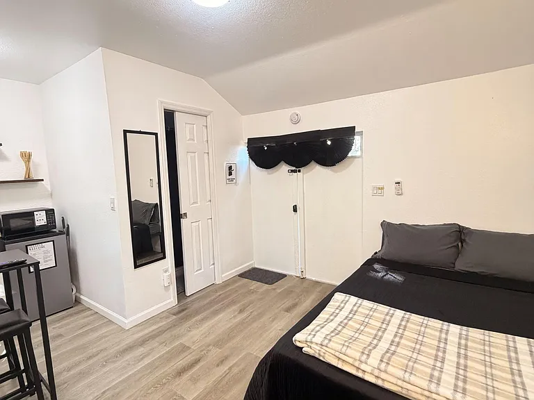 Furnished private bedroom with wood-look flooring, a full bed, mini-fridge and microwave, plus a closet and wall mirror.