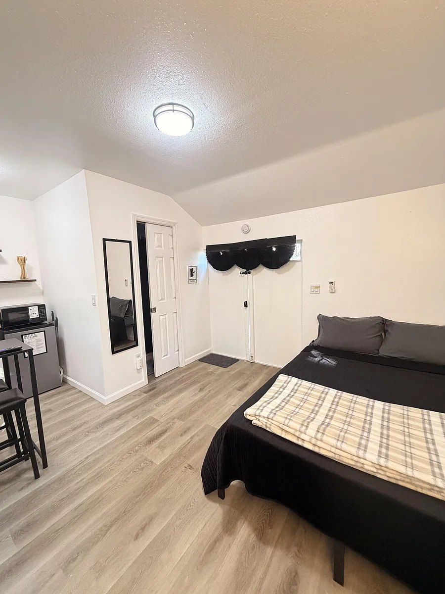 Furnished private bedroom with wood-look flooring, a full bed, mini-fridge and microwave, plus a closet and wall mirror. - May St