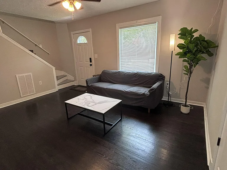 Open living area with dark wood-style flooring, ceiling fan, and a large window near the entry and stairway.