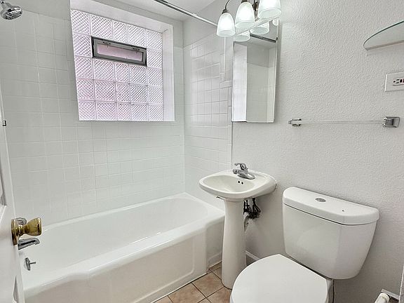 Bright and modern bathroom featuring a sleek bathtub, stylish pedestal sink, and an elegant lighting fixture, enhanced with natural light from a frosted window.