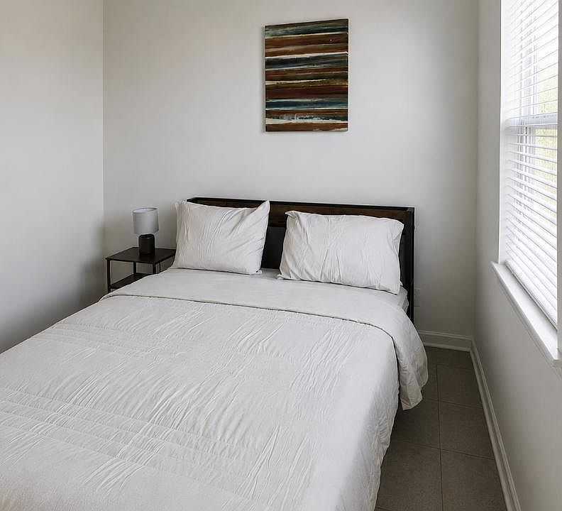 Private bedroom with a full bed, nightstand and lamp, plus a window with blinds for natural light and privacy.