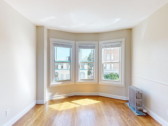 Bright, sunny corner of a room with three large bay windows, hardwood floor, and a vintage radiator against the wall.