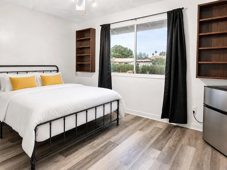 Private bedroom with a full bed, wide window with curtain panels, built-in wall shelving, and durable wood-look flooring.