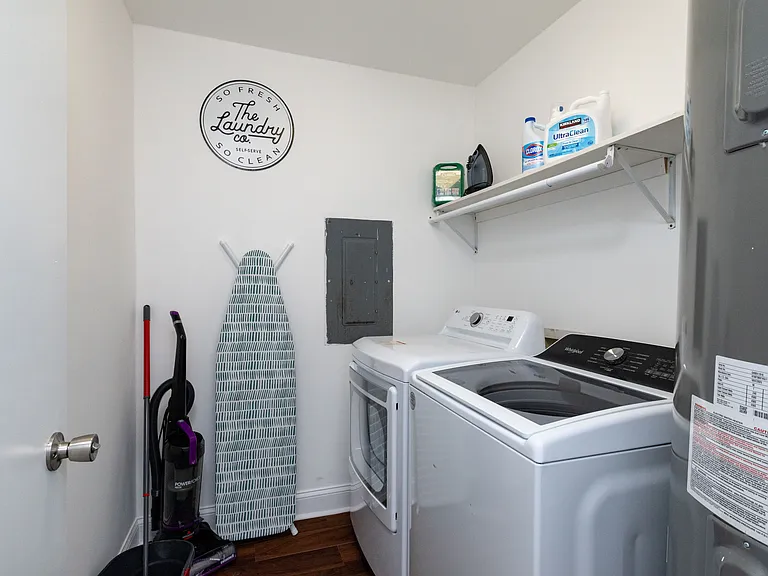 Convenient laundry room with washer, dryer, ironing board, and cleaning supplies.