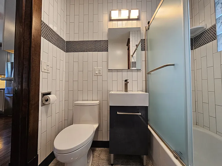 Bathroom is connected through the kitchen. Bathroom is fully tiled and includes a tub with enclosed glass surround, towel / storage area, under sink cabinet, and mirrored vanity.