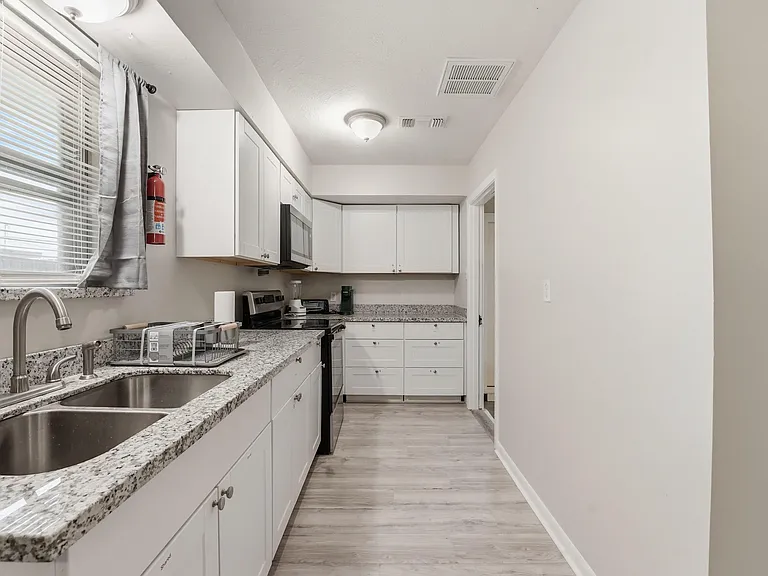 Galley-style kitchen with ample white cabinetry, granite-look counters, double sink, and black appliances for easy meal prep.
