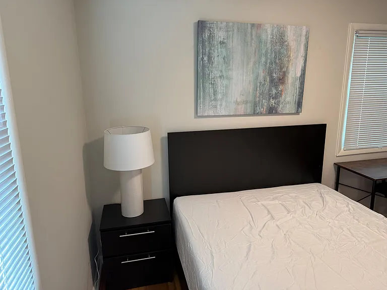 Furnished bedroom with a comfortable bed, nightstand and lamp, plus windows with blinds for adjustable natural light.