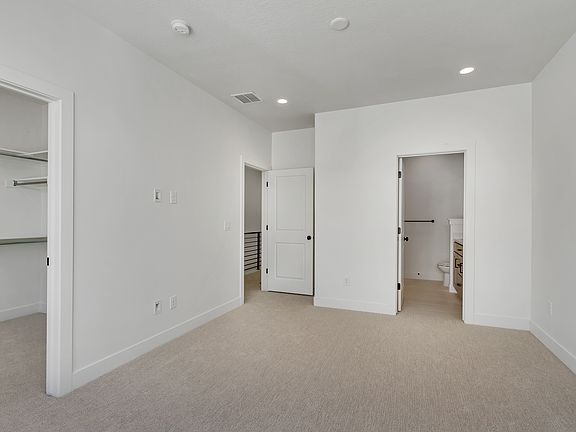 A minimalist empty room with beige carpet, white walls, a walk-in closet, and a door leading to a ba