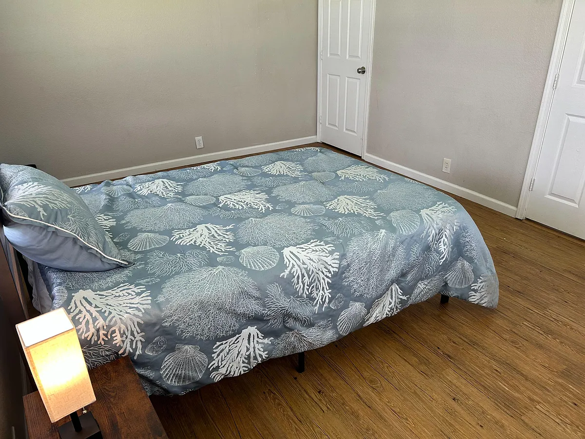 Furnished bedroom with wood-style flooring, neutral walls, and a bed setup with nearby nightstand and lamp. - Pecan St