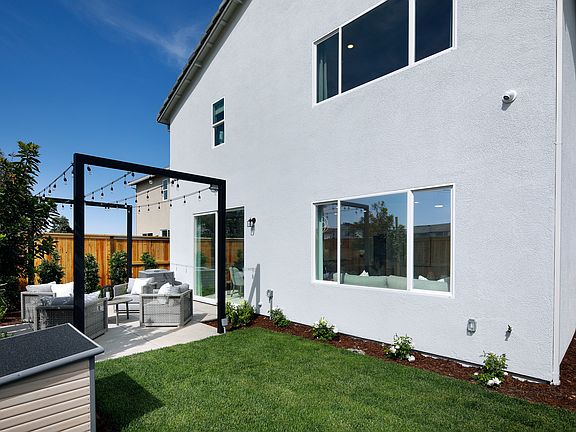 A modern, white two-story house with large windows and a covered patio area in the foreground, surro
