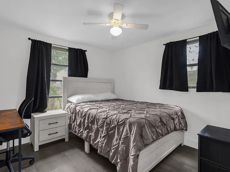 Furnished bedroom with a full bed, nightstand, and desk, plus ceiling fan and two windows with blackout curtains.