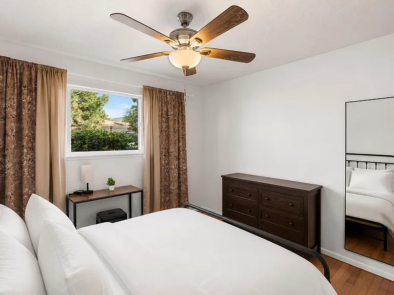 Furnished bedroom with a ceiling fan, large window with curtains, dresser storage, and a full-length mirror.