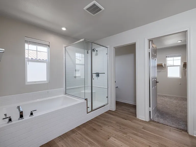 Spacious master bathroom showcasing access to the walk-in closet, glass-enclosed shower, soaking tub, and private toilet room. Bright natural light and laminate flooring create a modern, airy retreat within this elegant Menifee home.