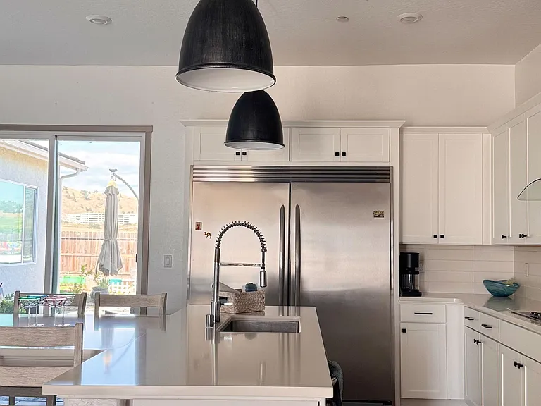 Kitchen features a large island with sink, ample white cabinetry, and stainless-steel refrigerator, plus pendant lighting.