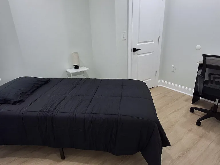 Furnished bedroom with a comfortable bed, nightstand, and dedicated desk setup, plus a window for natural light.