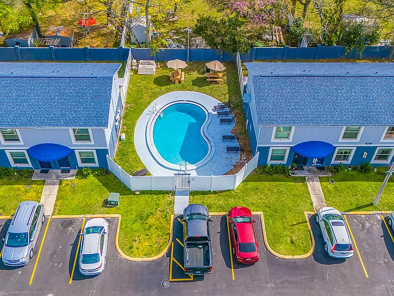 Charming aerial view of a residential complex featuring blue-roofed buildings, a kidney-shaped pool, lush green lawn, and ample parking.
