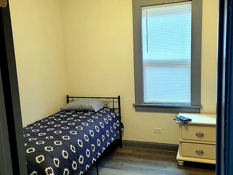 Furnished private bedroom with a twin bed, nightstand, ceiling fan, and a window with blinds for light control.